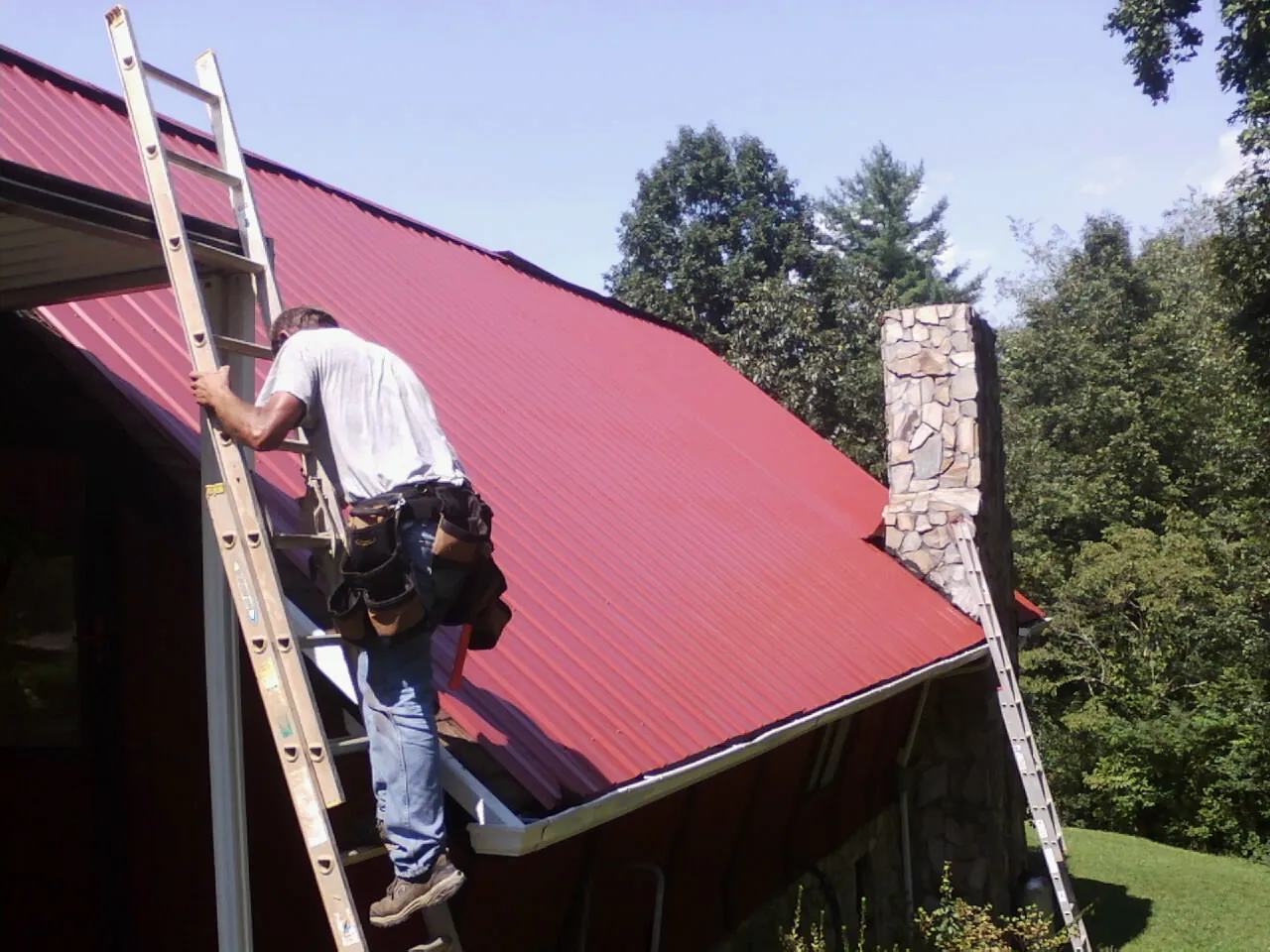 Roofer on a ladder beside a red metal roof and stone chimney — Soffit & Fascia Repair by Roofing Brothers in Fort Calhoun